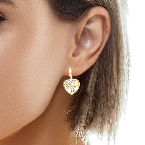 A close-up image showcases a woman's ear adorned with the "Tree of Life and Heart Dangle Earrings," meticulously crafted in 14k gold-filled material. The earrings elegantly dangle, partially concealed by her short hair, while the white background accentuates their intricate floral patterns and exquisite details.
